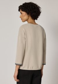 Frau mit lockigem Haar, die ein beige langärmeliges Oberteil mit schwarz-weiß gestreiften Bündchen trägt, blickt nach links vor einem schlichten Hintergrund.