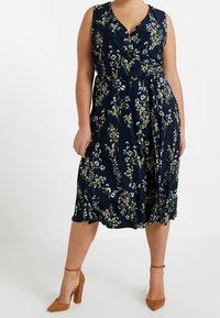 Blommig wrap-klänning i marinblått med V-ringning, ärmlös design, justerbar midja och knälång kjol. Har vita och gröna blommönster.