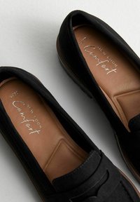 Black loafers with a smooth suede upper and a tan cushioned insole. Features a decorative strap and slight heel for support.