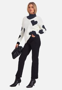 Weißer Strickpullover mit schwarzen Herzmustern, kombiniert mit schwarzen eng anliegenden Hosen und schwarzen h Camelot-Stiefeln, hält eine schwarze Clutch.