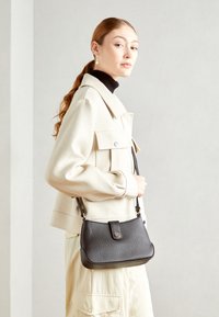 Czarna torebka na ramię z ziarnistej skóry o zaokrąglonym kształcie, z mosiężnymi okuciami i zamknięciem na górną klapę. Noszona z kremową kurtką i czarnym golfem.