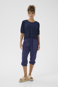 Blusa blu navy con inserti a occhio di pernice e maniche a sbuffo, abbinata a pantaloni larghi di un viola intenso e sandali intrecciati di un beige chiaro.