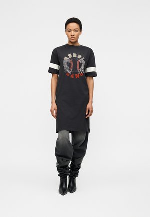 Femme portant une robe T-shirt noire oversize avec des rayures blanches aux manches, un dessin de tigre, le texte "Rebel Gang", un jean gris ample et des bottes noires décontractées.