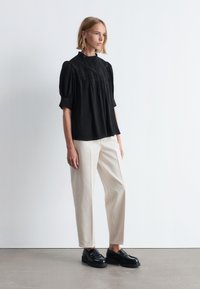 Schwarze, strukturierte Bluse mit Puffärmeln und hohem Kragen, kombiniert mit cremefarbenen Hosen und schwarzen klobigen Schuhen. Minimalistisches Design, neutrale Farben.