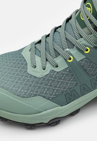 Scarpa da trekking verde con tomaia in mesh testurizzato, dettagli in plastica, lacci riflettenti e suola in gomma resistente per una migliore aderenza.