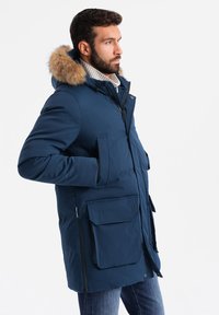 Marinblå parka-jacka med foder av päls i huvan, dragkedja och knappstängning, flera framfickor och en vadderad textur.