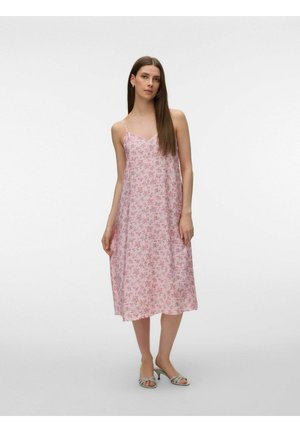Femme debout portant une robe midi rose à motifs floraux avec des bretelles fines et des sandales à talons clairs, sur un fond uni.