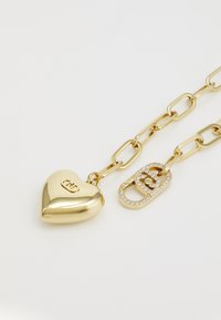 Gouden hartvormige hanger met reliëfdetails, verbonden met een gouden schakelkettinkje met een slotontwerp versierd met rhinestones.