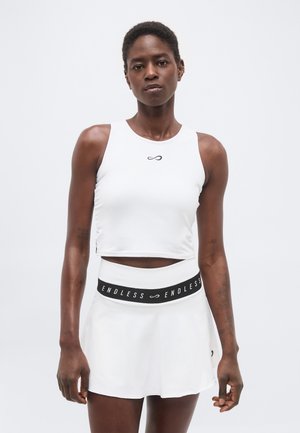 Αθλητική γυναίκα που φοράει λευκό αμάνικο crop top και λευκή φούστα με μαύρη μέση που γράφει "ENDLESS" σε απλό φόντο.