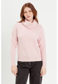 Pull à col roulé en tricot rose clair avec un ourlet et des poignets côtelés. Coupe légèrement oversize, texture douce. Porté avec un pantalon gris foncé.