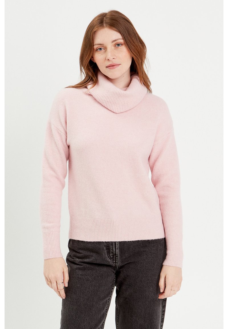 Pull à col roulé en tricot rose clair avec un ourlet et des poignets côtelés. Coupe légèrement oversize, texture douce. Porté avec un pantalon gris foncé.