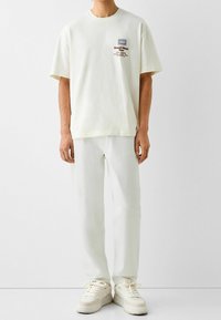 Camiseta oversize de color crema claro con estampado gráfico, combinada con pantalones rectos a juego y zapatillas blancas gruesas. Tejido de algodón casual.