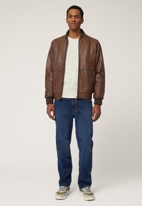 Braune Leder Bikerjacke mit gerippten Bündchen und Kragen, über einem strukturierten weißen Strickpullover getragen, kombiniert mit blauen Jeans und grauen Sneakers.