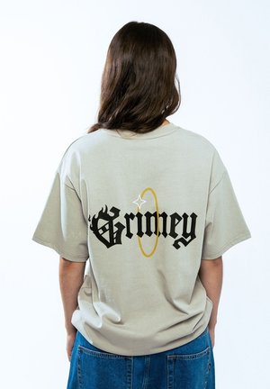 Camiseta de gran tamaño gris claro con letras en negrita negras "Grimey" y un diseño de círculo dorado en la parte posterior. La tela parece suave y tiene un ajuste relajado.