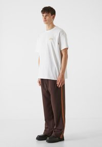 Bijela pamučna majica s malim zlatnim logotipom na prsima, uparen s smeškim trenirkama koje imaju narančaste i žute pruge duž bočnih strana.