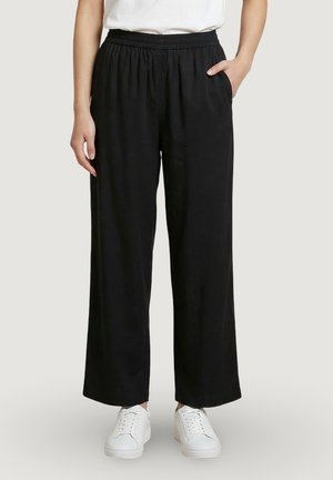 Žena v černých kalhotách s širokými nohavicemi a elastickým pasem a bílými teniskami, stojící s jednou rukou v kapse na neutrálním pozadí.