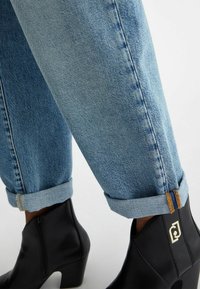 Vaaleansiniset farkut käärittyinä, yhdessä mustien nilkkaterien kanssa, joissa on kultainen symboli sivussa ja matala puolikeskijalka.