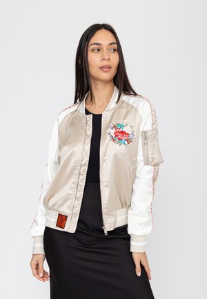Donna con capelli lunghi e scuri che indossa una giacca bomber in raso beige e bianca con ricami floreali e un vestito nero, che guarda di lato.