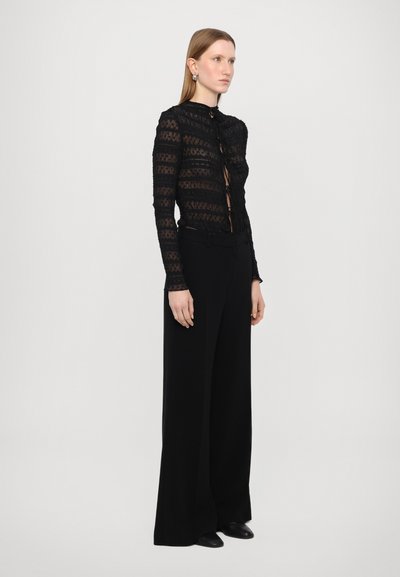 Zwarte sheer kanten top met strepen, hoge hals en voorste strikdetail, gecombineerd met zwarte wijde broeken en zwarte schoenen.