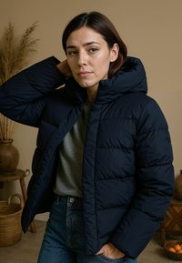 Marine blauwe gewatteerde jas met doorgestikte panelen en een capuchon, gecombineerd met een grijze top. De stof lijkt glad; het ontwerp is iets korter.