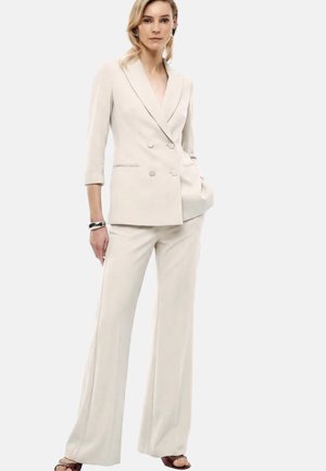 Donna che indossa un blazer doppiopetto color crema e pantaloni larghi coordinati, in piedi con una mano in tasca e un'espressione neutra.