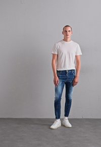 Man standing against gray wall wearing white t-shirt, blue paint-splattered skinny jeans, and white sneakers on concrete floor.