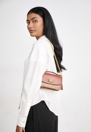 Woman with long black hair wears white shirt, black pants, and a brown leather and fabric shoulder bag with gold chain strap.