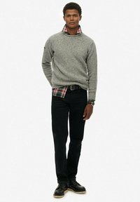 Suéter de punto gris, superpuesto sobre una camisa de cuadros roja y verde, combinado con jeans negros y zapatos negros de cordones. Diseño simple y texturizado.