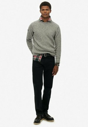 Suéter de punto gris, superpuesto sobre una camisa de cuadros roja y verde, combinado con jeans negros y zapatos negros de cordones. Diseño simple y texturizado.