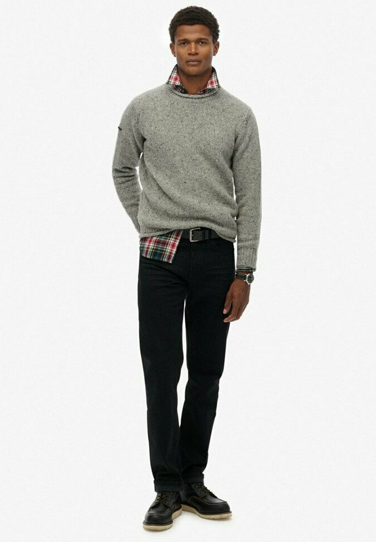 Suéter de punto gris, superpuesto sobre una camisa de cuadros roja y verde, combinado con jeans negros y zapatos negros de cordones. Diseño simple y texturizado.