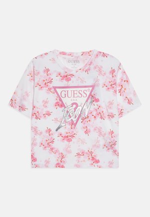 T-shirt blanc avec impression florale rose, manches courtes, col rond, présentant un logo GUESS triangulaire en rose et accents argentés.