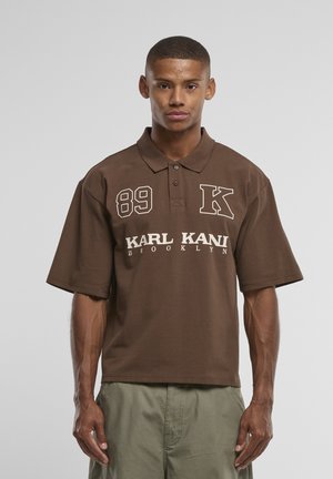 Noor mees seisab, tal on seljas pruun polosärk, millel on tekstid „89 K“ ja „Karl Kani Brooklyn“, ning helerohelised püksid, taust on ühevärviline.