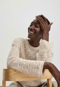 Cremig-weißer Strickpullover mit einer lockeren, offenen Maschenstruktur und gerippten Bündchen; Modell sitzt auf einem hellen Holzstuhl und zeigt glatte Haut und kurze Haare.