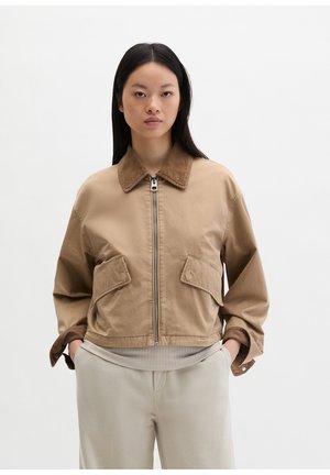 Kobieta o długich ciemnych włosach nosi beżową kurtkę na zamek z dużymi przednimi kieszeniami oraz pasujące spodnie, stojąc na tle jednolitego białego tła.