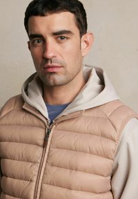 Kamizelka w kolorze jasnego beżu z pikowanym wzorem, z zamkiem błyskawicznym oraz beżową bluzą z kapturem pod spodem.