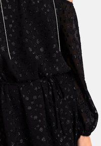 Robe noire en tissu texturé avec des points brillants surélevés, à longues manches transparentes, et une taille cintrée avec un détail de lien décoratif.