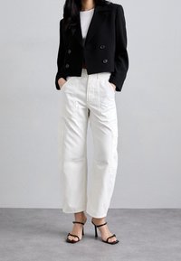 Mujer vestida con una chaqueta corta negra de doble breasted, pantalones cargo blancos de pierna ancha y sandalias de tacón alto negras, de pie sobre un suelo gris.