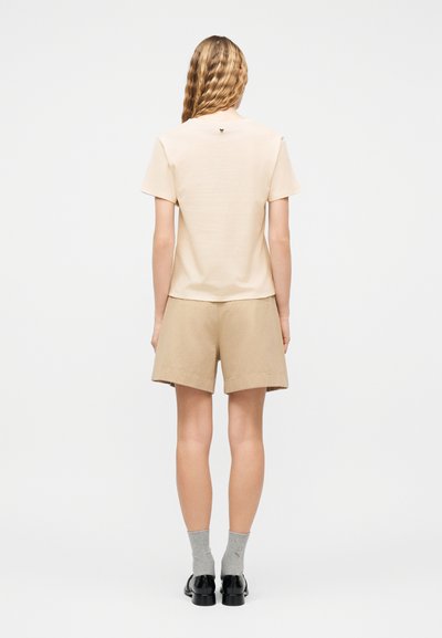 Γυναίκα με κυματιστά ξανθά μαλλιά, που φοράει μπεζ μπλουζάκι με κοντά μανίκια, ταιριαστά μπεζ σορτς, γκρι κάλτσες και μαύρα παπούτσια, στέκεται στραμμένη με την πλάτη σε λευκό φόντο.