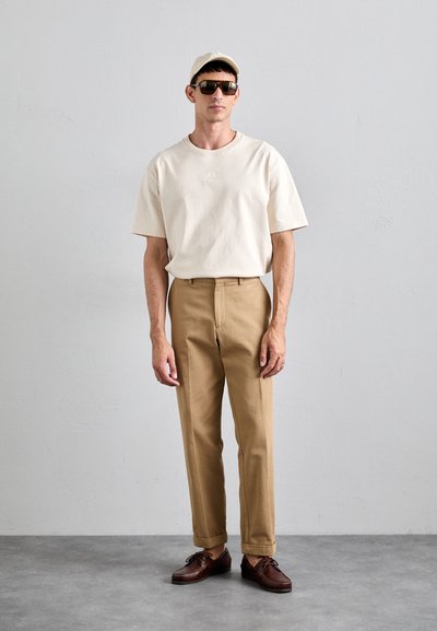 Beige bomull t-skjorte, lysebrune bukser, brune lærersko. En avslappet caps og mørke solbriller fullfører antrekket. Enkelt, moderne design.