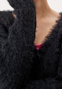 Černý huňatý cardigan s texturovaným, nadýchaným povrchem, s hlubokým véčkovým výstřihem a jedním černým knoflíkem na zapínání. Růžová spodní vrstva je viditelná.