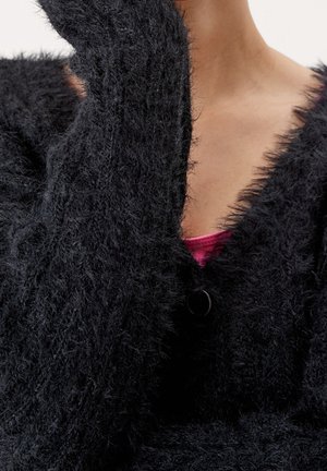 Czarny, puszysty kardigan o teksturowej, fluffy powierzchni, z głębokim dekoltem w kształcie litery V i pojedynczym zapięciem na czarny guzik. Widoczna różowa warstwa podstawowa.