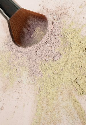 Makeup brush with brown bristles lying on scattered pastel-colored powders, featuring purple and yellow hues against a light background.