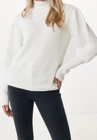 Pull col roulé blanc en maille avec poignets côtelés et motifs texturés, associé à un jean gris foncé, créant un look en couches.