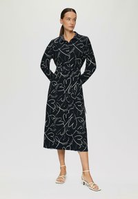 Lungo vestito nero con motivi bianchi a forma di cuore, design con colletto, vita con cintura e maniche lunghe. Completato da sandali bianchi con cinturini.