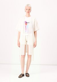 Cardigan alb tricotat, cu mâneci scurte, având un model texturat. Purtați peste un tricou grafic și pantaloni scurți din dantelă, asortat cu pantofi cu imprimeu animalier.