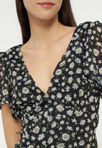 Femme portant une robe noire à motif floral marguerite blanche avec un décolleté en V, montrant le décolleté ainsi qu'une partie du visage et un tatouage sur le bras.