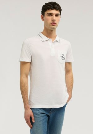 Hombre joven que lleva una camisa polo blanca con ribete negro en el cuello y un pequeño logo en el bolsillo del pecho, combinada con pantalones vaqueros azules.