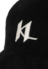 Fekete kordbársony sapka, amelyen elöl fehér hímzett "KL" logó található. A textúra bordás, ívelt karimával rendelkezik.