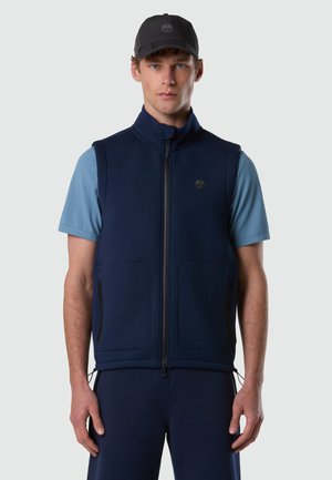 Junger Mann mit schwarzer Kappe, hellblauem Kurzarmhemd und marineblauem Reißverschlussweste mit Taschen, steht vor einem einfarbigen Hintergrund.