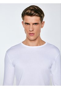 Jauns vīrietis ar īsiem brūniem matiem, valkājot vienkāršu baltu krekla ar garām rokām, skatās tieši priekšā ar neitrālu izteiksmi.